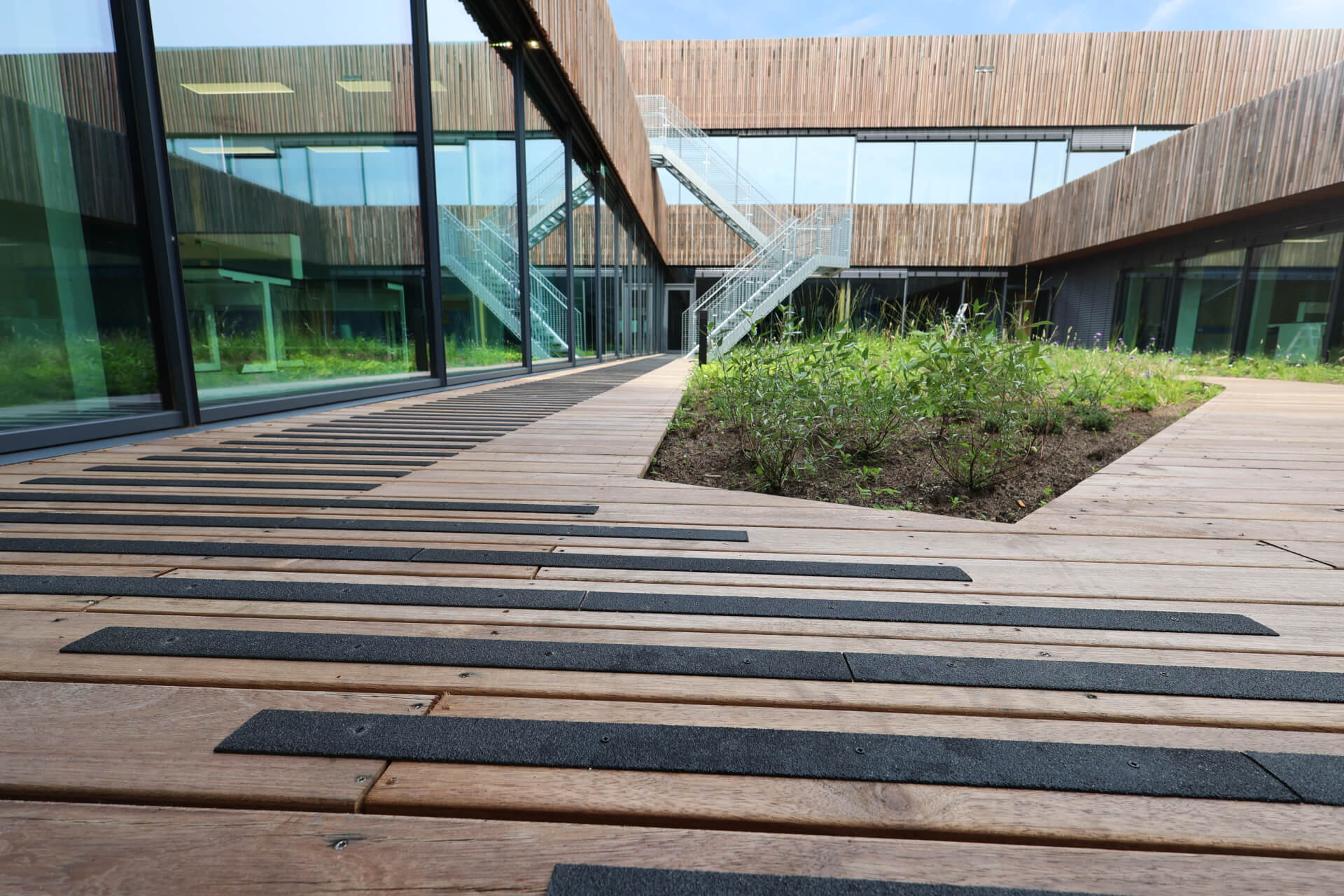 Vlonder antislip maken door Vlonderhoud Hardhouten vlonder met zwarte antislip-strips langs een moderne glazen gevel — vlonder antislip maken door Vlonderhoud