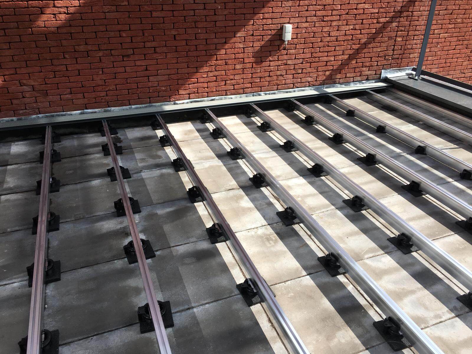 Bevestigingsmateriaal vlonder — Vlonderhoud Aluminium onderconstructie op verstelbare tegeldragers en rubberen afstandhouders op een betonnen terras langs een bakstenen muur — bevestigingsmateriaal vlonder van Vlonderhoud
